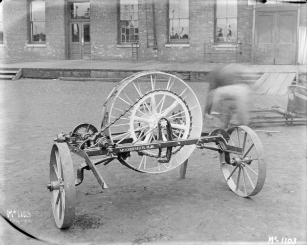 Wheels and chain drive of binders outdoors at McCormick Works. A loading dock and brick factory building are in the background. Two men, (blurred by motion) are working behind the binder on the right.