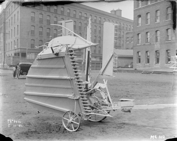 Reaper with bed folded up outdoors at McCormick Works. In the background are railroad cars, an automobile, and brick factory buildings.