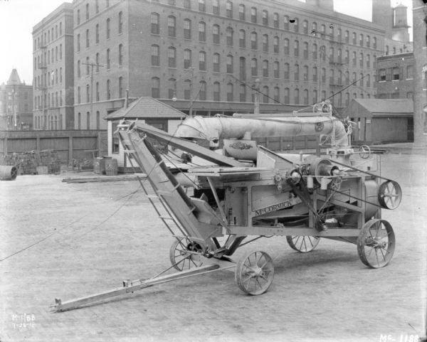 McCormick Thresher outdoors in the yard at McCormick Works. In the background are brick factory buildings and a tall, wooden fence.