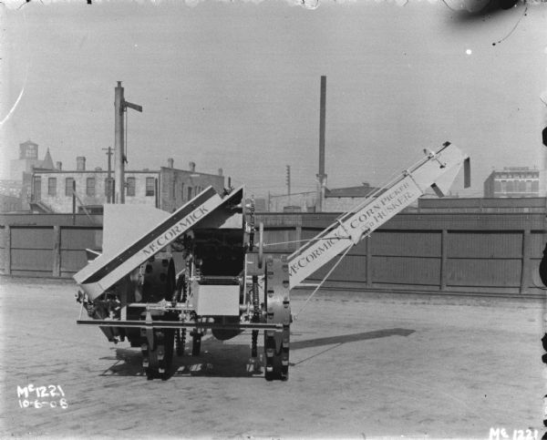 McCormick Corn Picker and Husker outdoors in a yard at the factory. In the background is a tall, wooden fence, and buildings beyond.