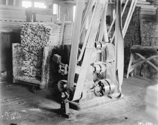 Manufacturing machine indoors on factory floor. There are wooden dowels stacked in a cart behind the belt-driven machine.