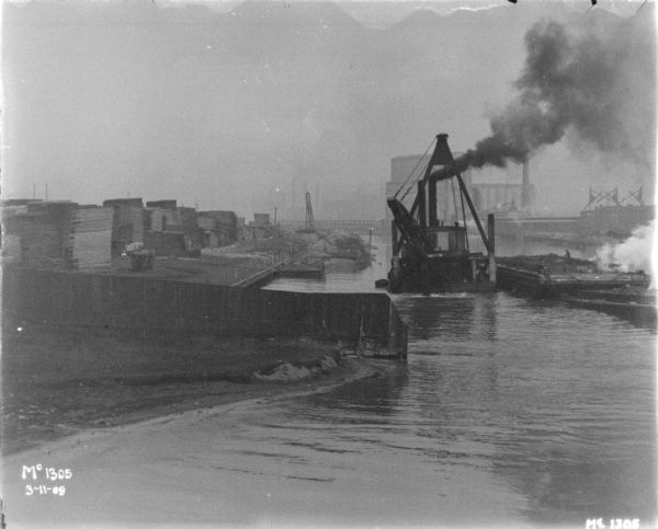Elevated view across water towards ships at the docks at McCormick Works. Lumber is stacked along the shoreline on the left.