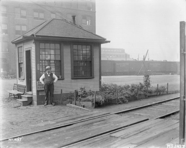 View across railroad tracks towards a man standing in front of a small building. There are plants in a fenced bed around the building. In the background are factory buildings and railroad cars.