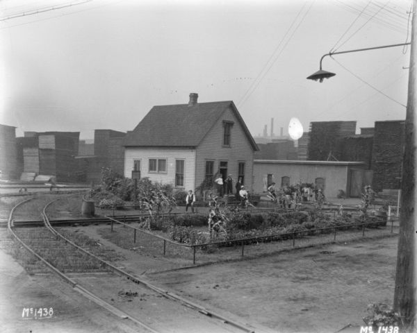 Elevated view across railroad tracks towards men working in a garden near a building at McCormick Works. There are tall stacks of lumber in the background.