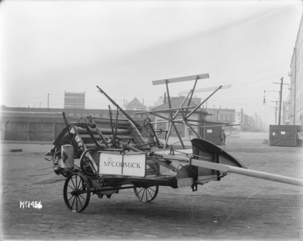 McCormick Binder outdoors in the yard at McCormick Works. In the background is a small building, with men standing near the gate, and a tall, wooden fence, with railroad cars behind the fence. Buildings are in the distance.