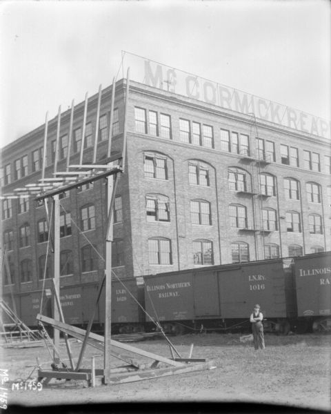 Hay stacker with arm raised in air outdoors in a yard at McCormick Works. A man is standing on the right, and behind him are railroad cars on railroad tracks, and a brick factory building. A large sign on top of the factory building reads, in part: "McCormick Reap"(cut off).