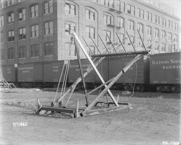 Hay stacker with lifter raised. In the background are railroad boxcars and a factory building.
