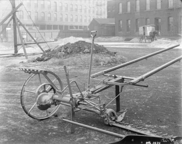 Mower outdoors in the yard at McCormick Works. In the background on the left is a man standing near a tall, wooden fence. Brick buildings are beyond the fence. In the background on the right is a horse-drawn vehicle near another brick factory building.