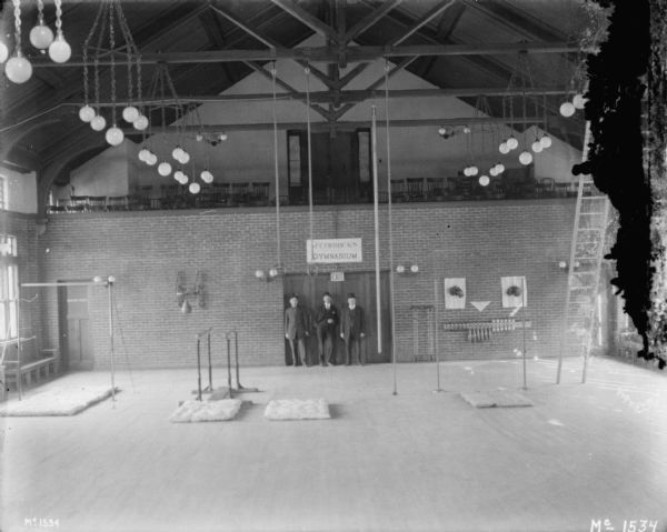 Interior, elevated view of McCormick's Gymnasium. Sports equipment is set up on the floor, and a punching bag is attached to the brick wall near a set of double doors. There is a balcony in the background lined with chairs. There are three men posing in front of the double doors. 