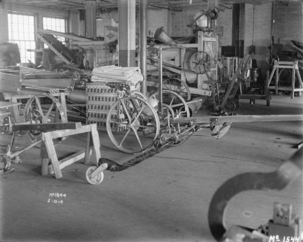 Mower indoors on factory floor. Other agricultural implements and parts are distributed throughout the room.