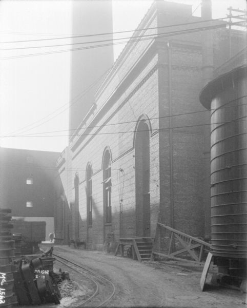 Railroad tracks along the side of a brick factory building at McCormick Works. A smokestack rises above the building in the background. There is a storage tank in the right foreground.