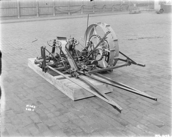 Corn Binder set-up on a pallet on the cobblestones outdoors in the yard at McCormick Works. In the background is a tall, wooden fence, and a small, wooden building on the far right.