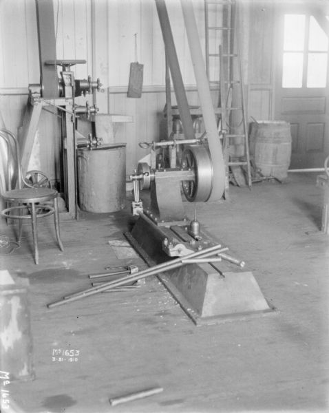 Indoor view of manufacturing and maintenance area in a factory building. A belt-driven machine is in the center, with a chair on the left. In the background on the right is a door to the outdoors.