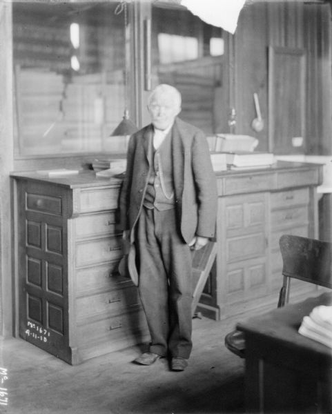 Full-length portrait of a man wearing a suit standing and posing by a large desk in an office.