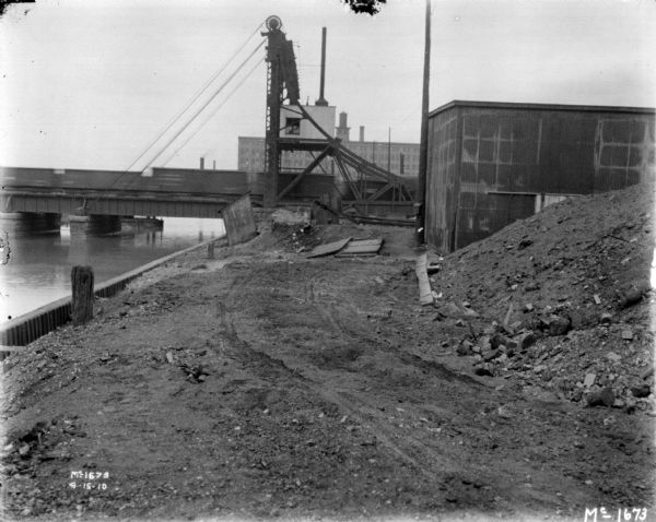 View along river of a yard at McCormick Works. There is a railroad train on a bridge across the river.