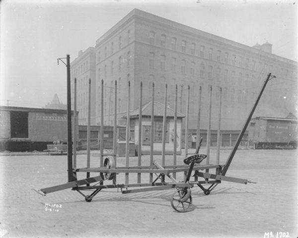 Sweep Rake outdoors in the yard at McCormick Works. In the background is a small, wooden sided building. In the background a man is working on the left near a railroad car. Behind the railroad car is a tall, wooden fence, and brick buildings are beyond.