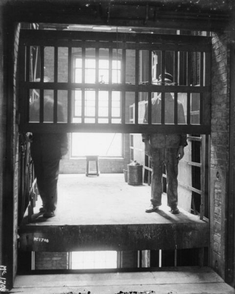 Two men riding freight elevator between floors in a factory. There is a window in the background.