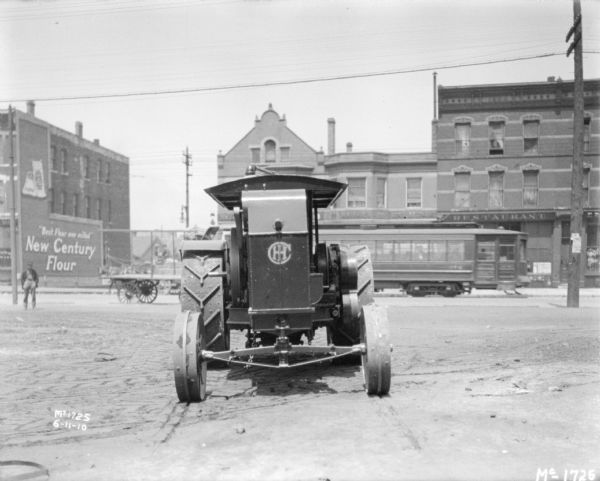 View towards the front of a tractor parked outdoors. In the background a man is standing along a street with a streetcar moving in front of storefronts.
