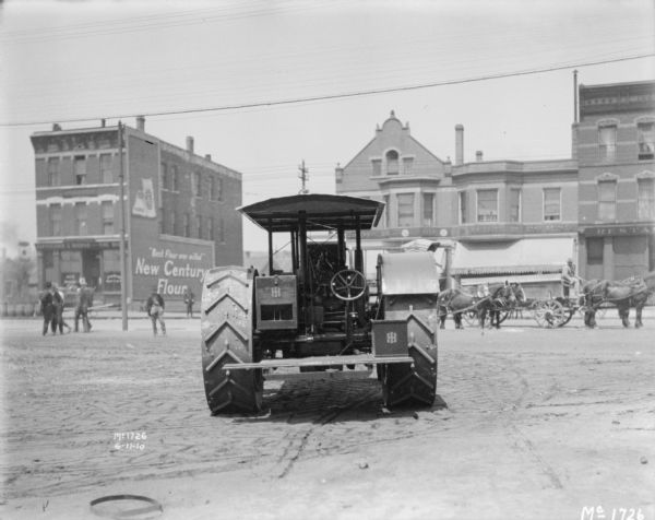 Rear view of a tractor at McCormick Works. In the background men are standing on the left near a street, with horse-drawn vehicles moving along the curb on the right near storefronts.