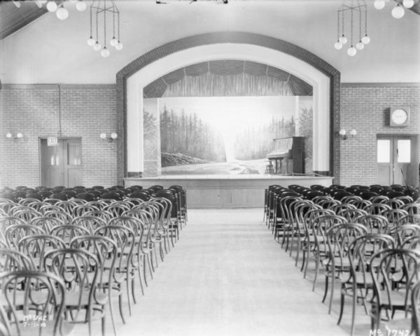 View of an auditorium with a stage. There is a mural painting at the back of the stage, and a piano with a stool is set up on the stage. Chairs are set up in rows on the floor.