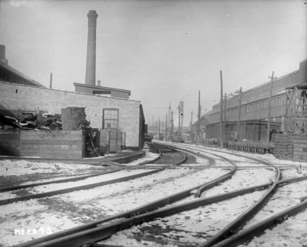 View across railroad tracks towards factory buildings. Snow is on the ground.