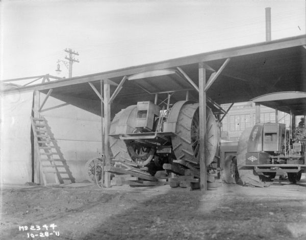 Tractor in an open-sided garage, with the back wheels propped on wood ramps. Another tractor is parked on the right.