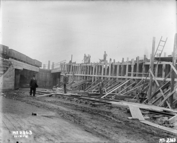 Men, some wearing suits, and other work clothes, are standing around a building under construction. There are railroad cars on a bridge on the left.