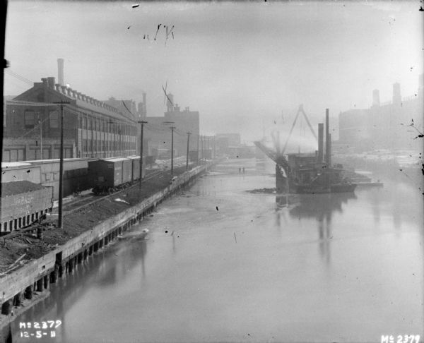 Elevated view of a ship, with a dredge, in a harbor. There are railroad cars on tracks near factory buildings on the left side of the harbor. There are more factory buildings on the right.