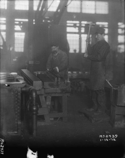 Man forming metal inside a factory building. A man is standing on the right holding up a sledgehammer.
