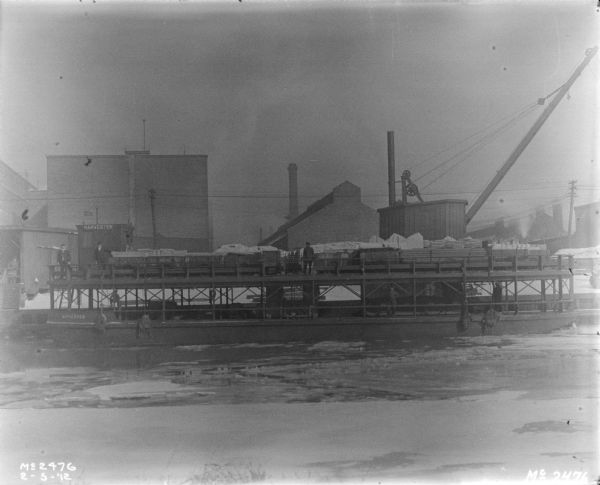 View across icy water towards men posing on a ship at a shipping pier at McCormick Works. Factory buildings are in the background.