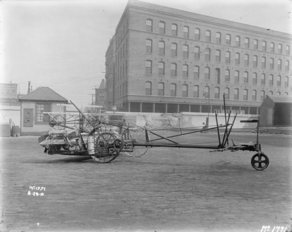 Push Binder outdoors in the factory yard at McCormick Works. In the background is a small, wooden building near a tall, wooden fence. Behind the fence are brick buildings. A man is sitting at the base of a lamppost in the background on the right.