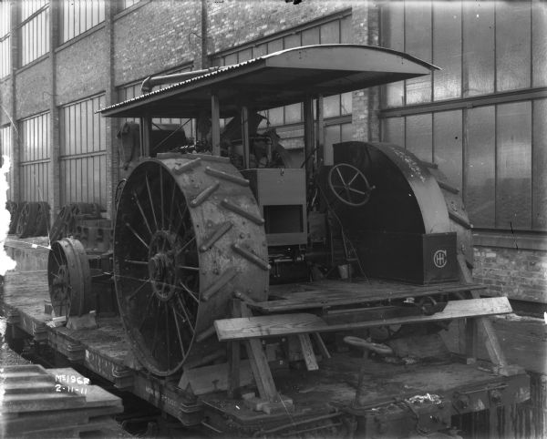 Tractor on flatbed railroad car for delivery at McCormick Works. A brick factory building is in the background.