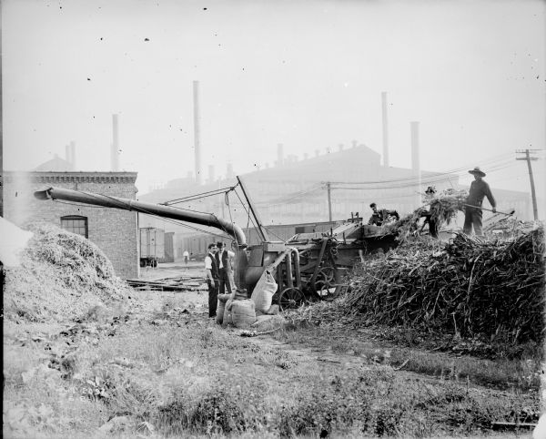 A group of men are operating a thresher in a plant yard at McCormick Works. Railroad cars and factory buildings are in the background.