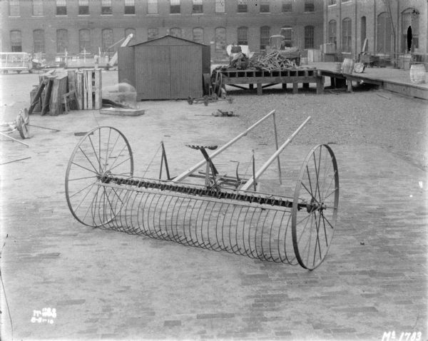 Dump rake in the yard at McCormick Works. There are pallets and other parts of agricultural machinery near a small building behind the dump rake. On the right is a loading dock near the side of a factory building. In the far background a man is working with other machinery in front of a large, brick factory building.