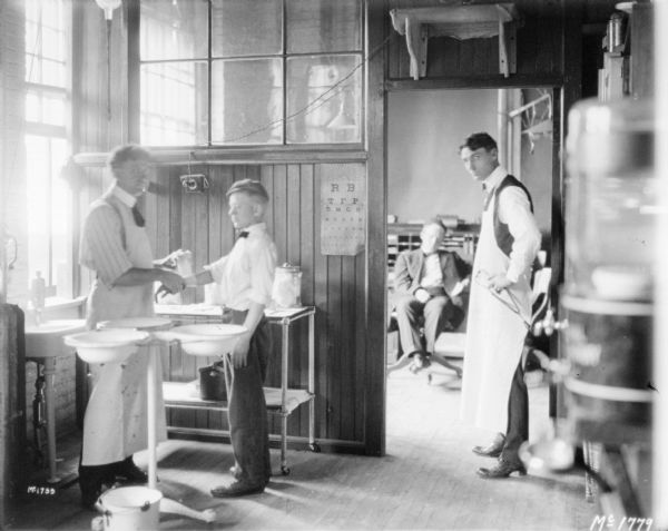A man wearing an apron is standing on the left near a sink while treating a boy's arm. Another man, also wearing an apron, is standing near an open doorway on the right, and through the doorway a man is sitting in a chair.