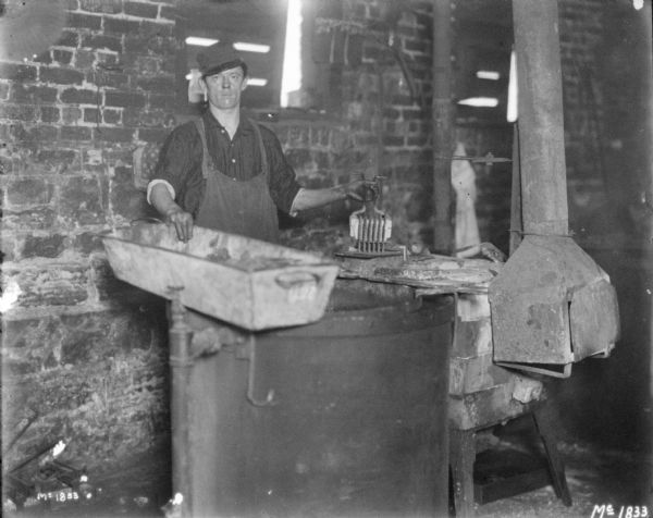 A man is standing in a forging area at McCormick Works. He is holding a long tub with his right hand, and holding another tool with his left hand.