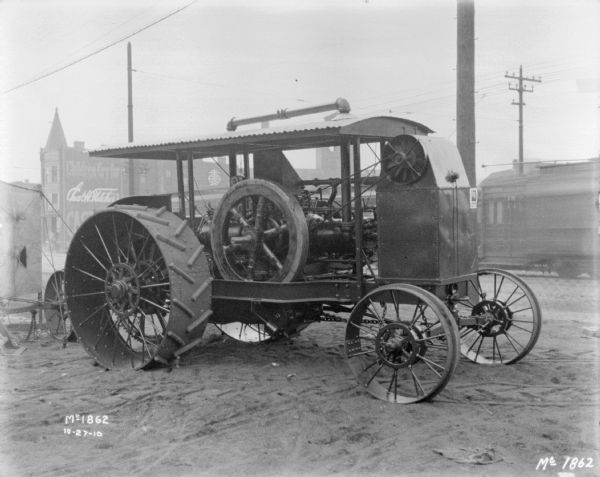 Three-quarter view from front right of a tractor in the yard at McCormick Works. There are commercial buildings and a streetcar along a street in the background. A sign on the side of a building reads: "Chas. H. Hetcher's."