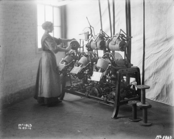 A woman is standing at a twine mill near the corner of a room, near a brick window. A white backdrop is set up behind the twine mill.