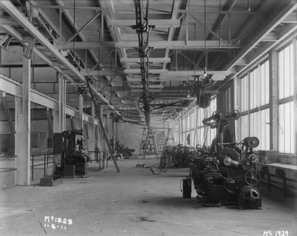Manufacturing area in a factory. A ladder is propped up against a beam near the ceiling. Windows line the wall along the right. Machinery is belt-driven.