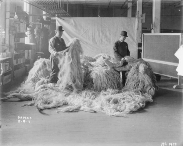 Men inspecting piles of raw sisal indoors at McCormick Works. A white backdrop is hanging in the background.