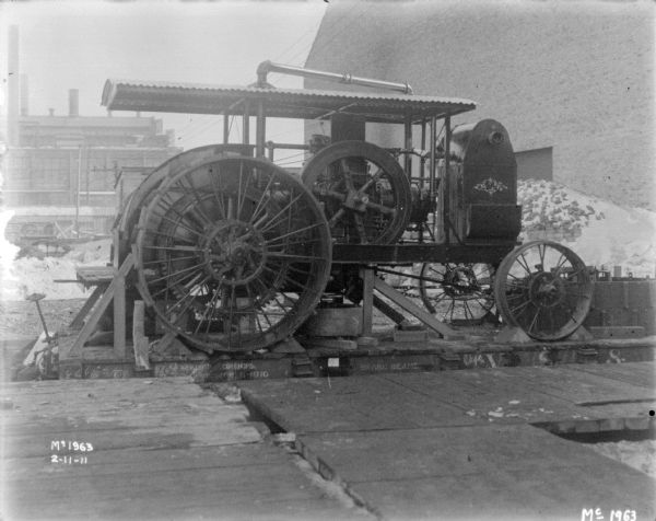 A tractor is positioned on a flatbed railroad car for delivery outdoors at McCormick Works. Factory buildings are in the background, and snow is on the ground.