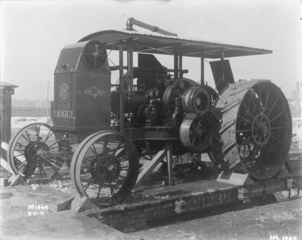 A Mogul tractor is positioned on a flatbed railroad car for delivery outdoors at McCormick Works. Factory buildings are in the background, and snow is on the ground.