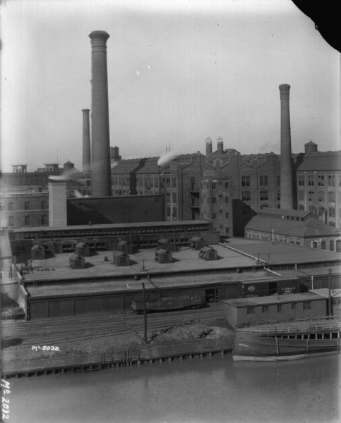 Aerial view across river towards McCormick Works, including factory buildings, smokestacks and a railroad yard. A ship named <i>Imperial</i> is along the dock in the right foreground.