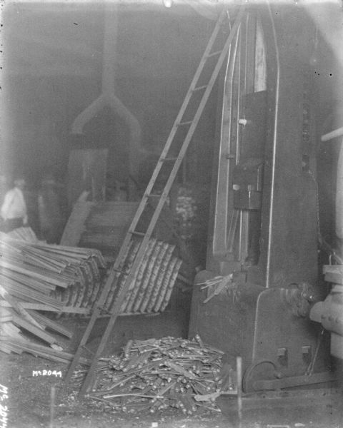 A ladder is leaning against the side of a manufacturing machine in the foreground, with machine parts in pile at the base. A man is standing in the background on the left.