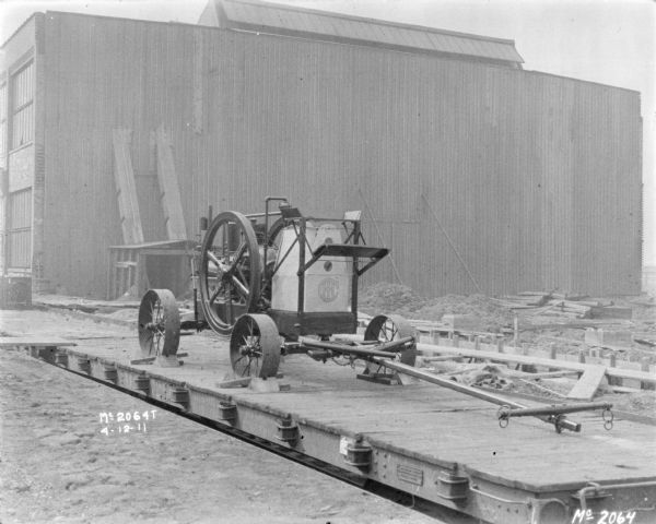 Tractor on a flatbed railroad car. A factory building is in the background.