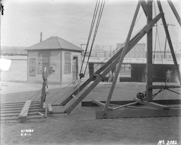 Hay Stacker outdoors in the yard at McCormick Works. In the background a man is standing in front of a small building, and other men are standing near an elevated railroad.