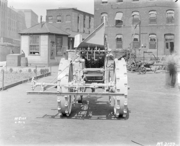 Binder outdoors in the yard at McCormick Works. Just behind the Binder on the left is a small, fenced garden near a small, wooden-sided building. In the distance are railroad cars on an elevated platform, and brick factory buildings. A man (blurred with motion) is walking on the right.