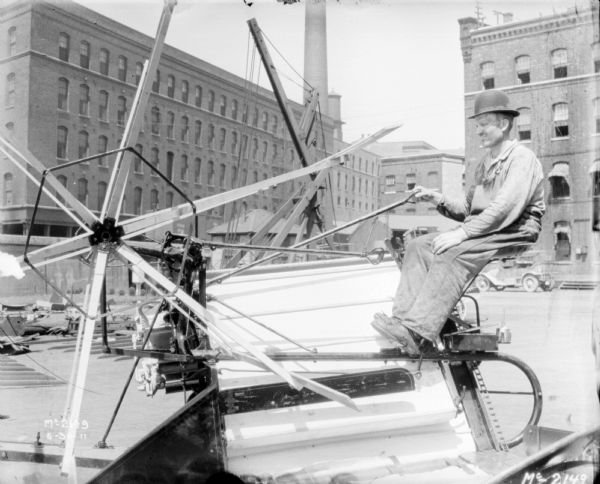 Close-up of a man riding a binder in the yard. Brick factory buildings are in the background.