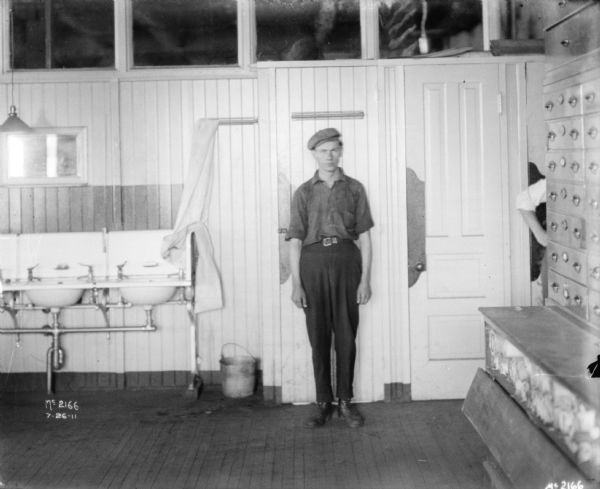 Male employee posing standing in a washroom at McCormick Works. A row of sinks are along the left. On the right a man (out of frame except for his arm) is standing behind a cabinet along the right wall.
