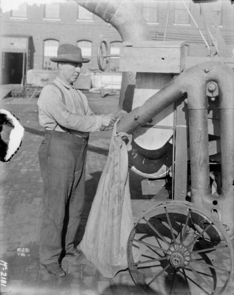 Man attaching sack to pipe of thresher outdoors in the yard at McCormick Works. In the background is a brick factory building.